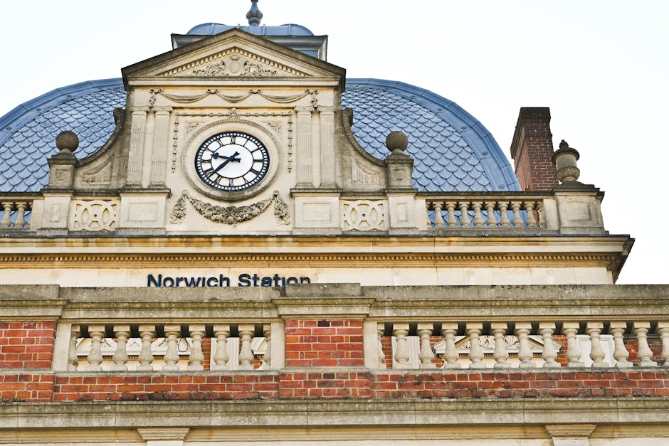 Close-up of the exterior of Norwich Station, featuring an ornate clock with black Roman numerals and hands set within a decorative stone frame. The building showcases classic architectural details, in